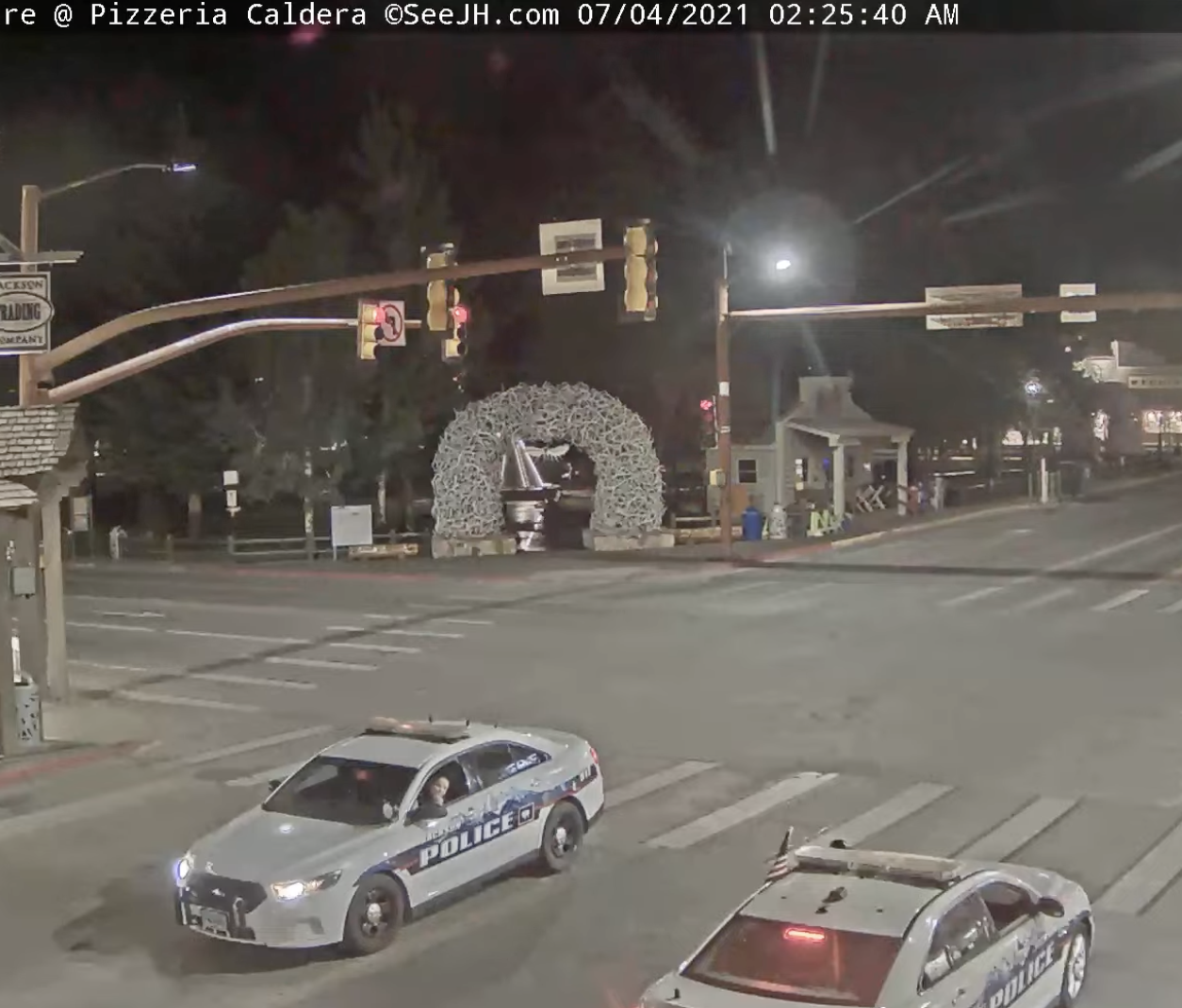A crop of a frame of a traffic cam timestamped 7/4/2021 2:25am. Two cop cars, driving opposite directions, have stopped in the middle of the intersection and rolled down their windows to talk to each other.