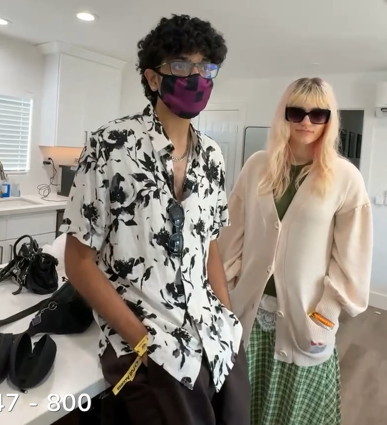 A young man with a Minecraft-style black and purple face mask, earrings, a silver chain, a black and white patterned button up shirt with the top buttons open and sunglasses hanging off the shirt, leaning back against the counter. On his right a young woman with pink and blonde hair, huge square rimmed purple sunglasses (even though they're indoors), wearing a cream sweater over a green top and a green skirt.