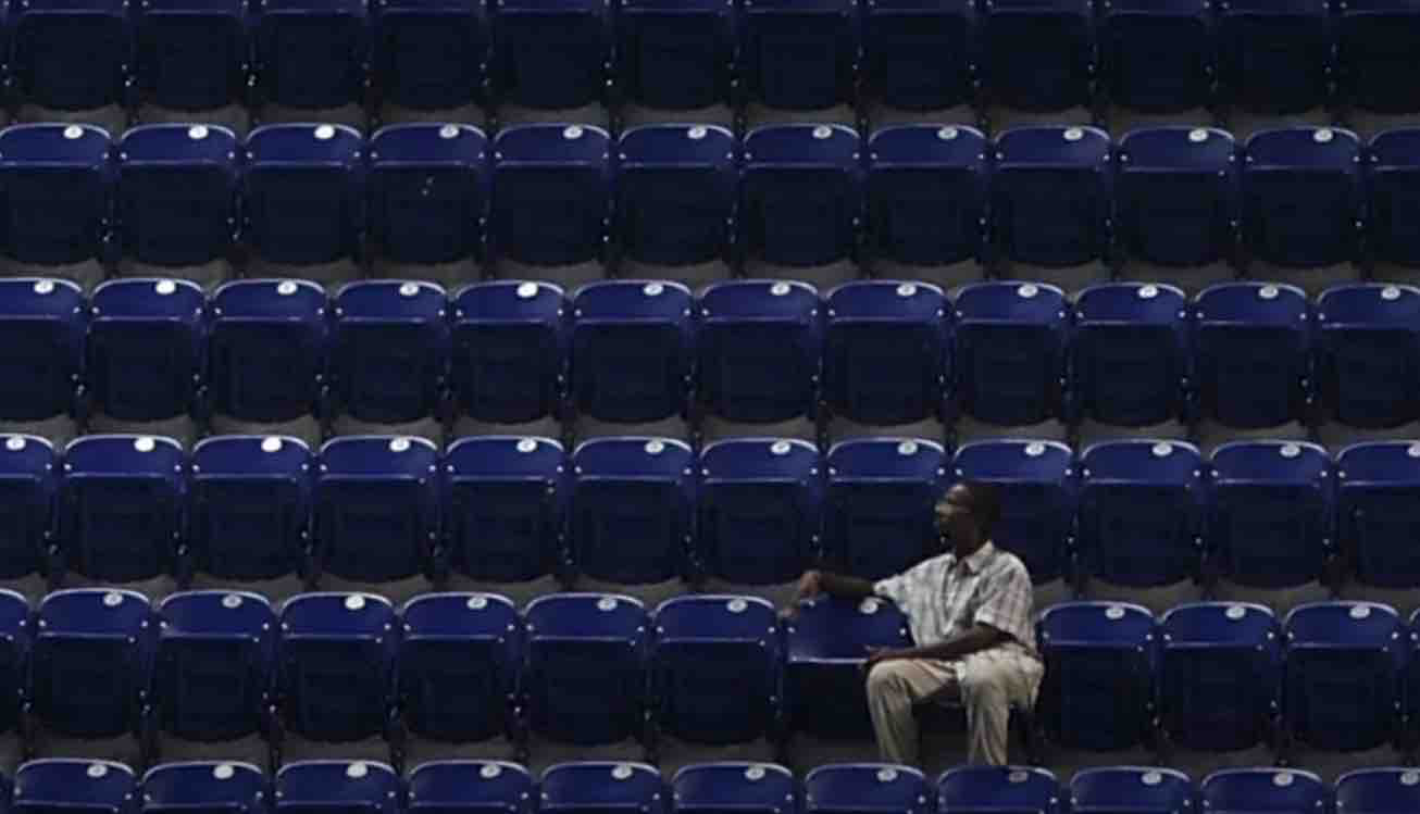 An older black man sits alone in a stadium. His face and body are angled sideways; he's not looking at the field.